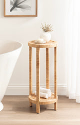 Tall round side table made of woven natural rattan with four vertical legs and a lower circular shelf, styled with a white vase and towels in a bright room.