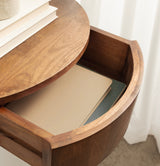 Close-up of round wooden side table with slid-open top revealing storage containing two books, showing smooth wood grain and curved edges.