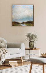 Softly colored landscape painting with a cloudy sky and reflective water, framed in wood, hanging above a beige sofa and side table with a glass vase of foliage.