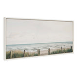 Framed canvas artwork depicting a tranquil beach scene with soft pale sky, calm ocean waves, sand, and a rope fence with grassy dunes in the foreground.