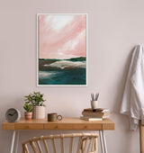 Abstract landscape painting with pink sky and green-blue water and hills, framed in white and hung above a wooden desk with plants, books, and a cup.