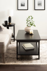 Rectangular black coffee table with lower shelf holding books, topped with a beige textured vase of greenery and a lit candle, set on patterned rug in neutral living room.