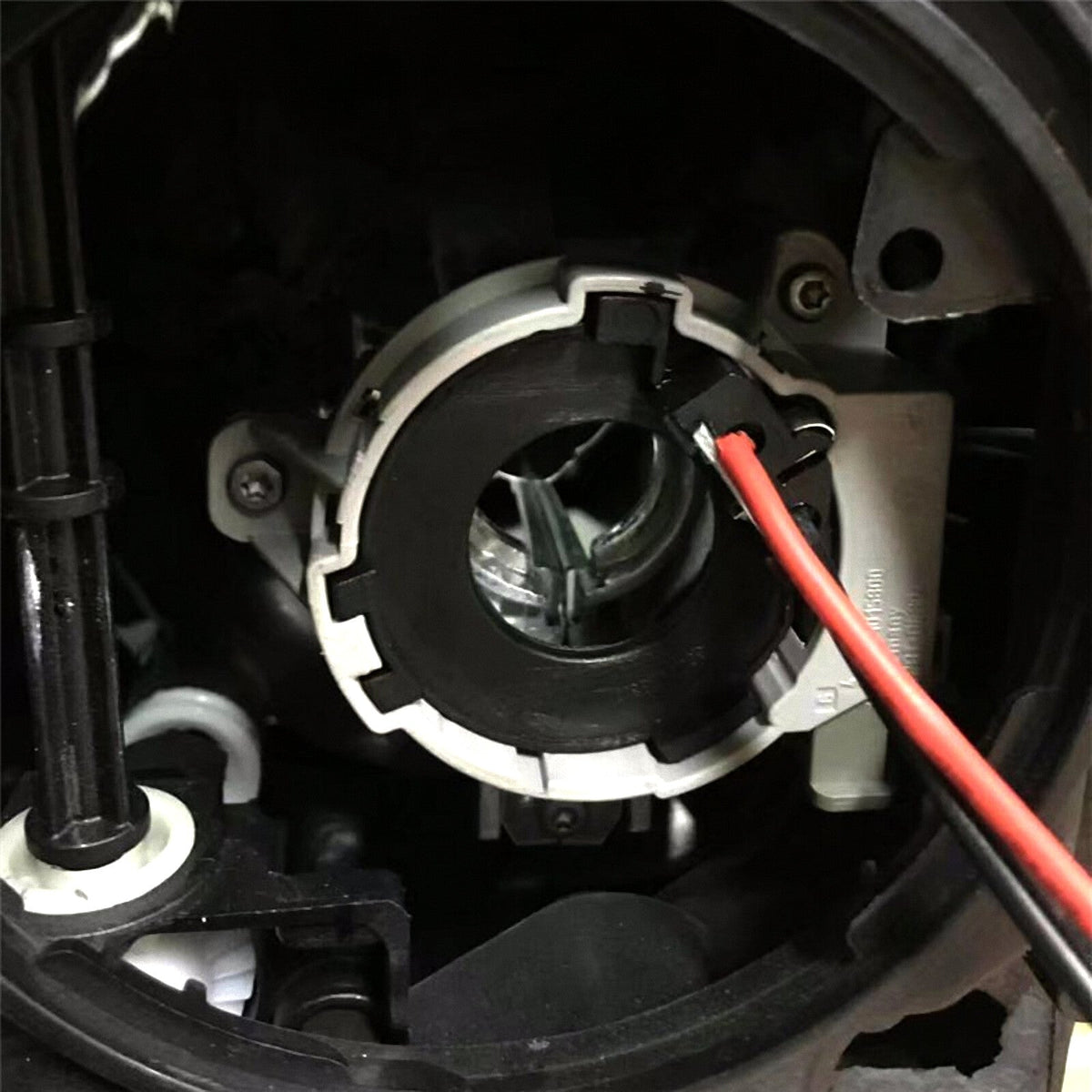 Close-up of a circular black and gray automotive headlight adapter with a red wire attached, mounted inside a vehicle's headlamp housing with surrounding metal and plastic parts.