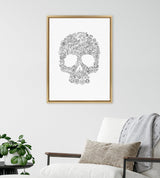 Framed black and white floral skull print by Viola Kreczmer hanging above a white armchair with gray throw and beige pillow, next to a green potted plant.