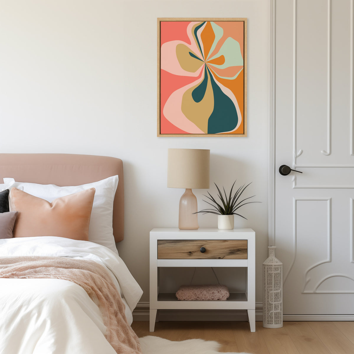 Abstract floral artwork in shades of coral, pink, teal, and mustard framed above a bedside table with a beige lamp and small spiky green plant, next to a bed with light peach and white bedding.