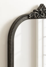 Close-up of an arched dark wood mirror frame featuring carved bead detailing along the inner edge and ornate leaf motifs atop the arch.