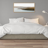Framed landscape photograph of a calm coast with hills under soft natural light, hung above a neatly made bed with white bedding and gray walls.
