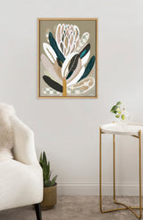 Abstract protea flower artwork with muted greens, browns, and beige tones in a light wooden frame above a modern gold and white side table with a small vase and plant.