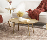 Oval Alessia coffee table with scalloped edges and gold finish, featuring tapered legs, topped with a small plant and stacked books, set on a patterned rug beside beige sofas with rust-colored knitted throws.