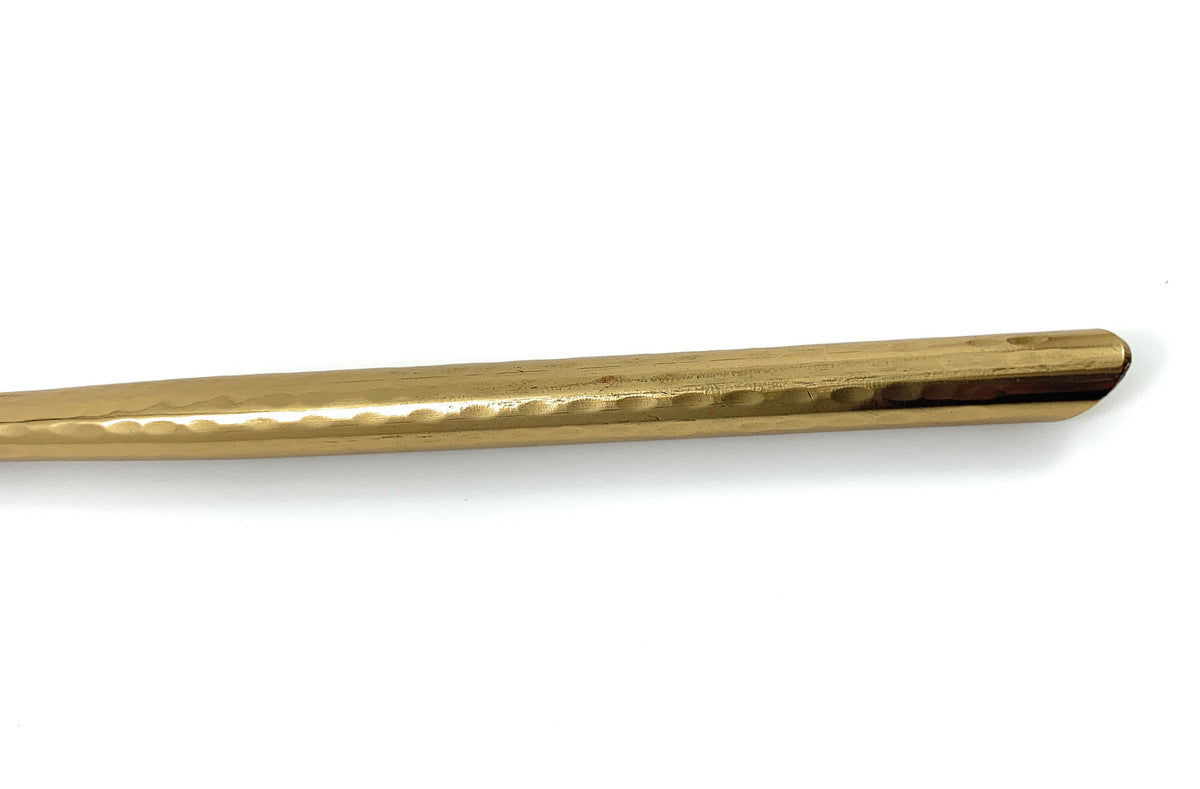 Close-up of the handle of a golden teaspoon with a hammered texture and smooth, rounded end against a white background.