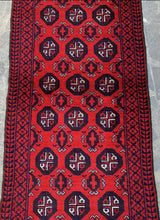 Hand knotted Afghani wool Baluch runner rug in red with intricate black geometric medallions and a border featuring repeating angular patterns, displayed on stone flooring.