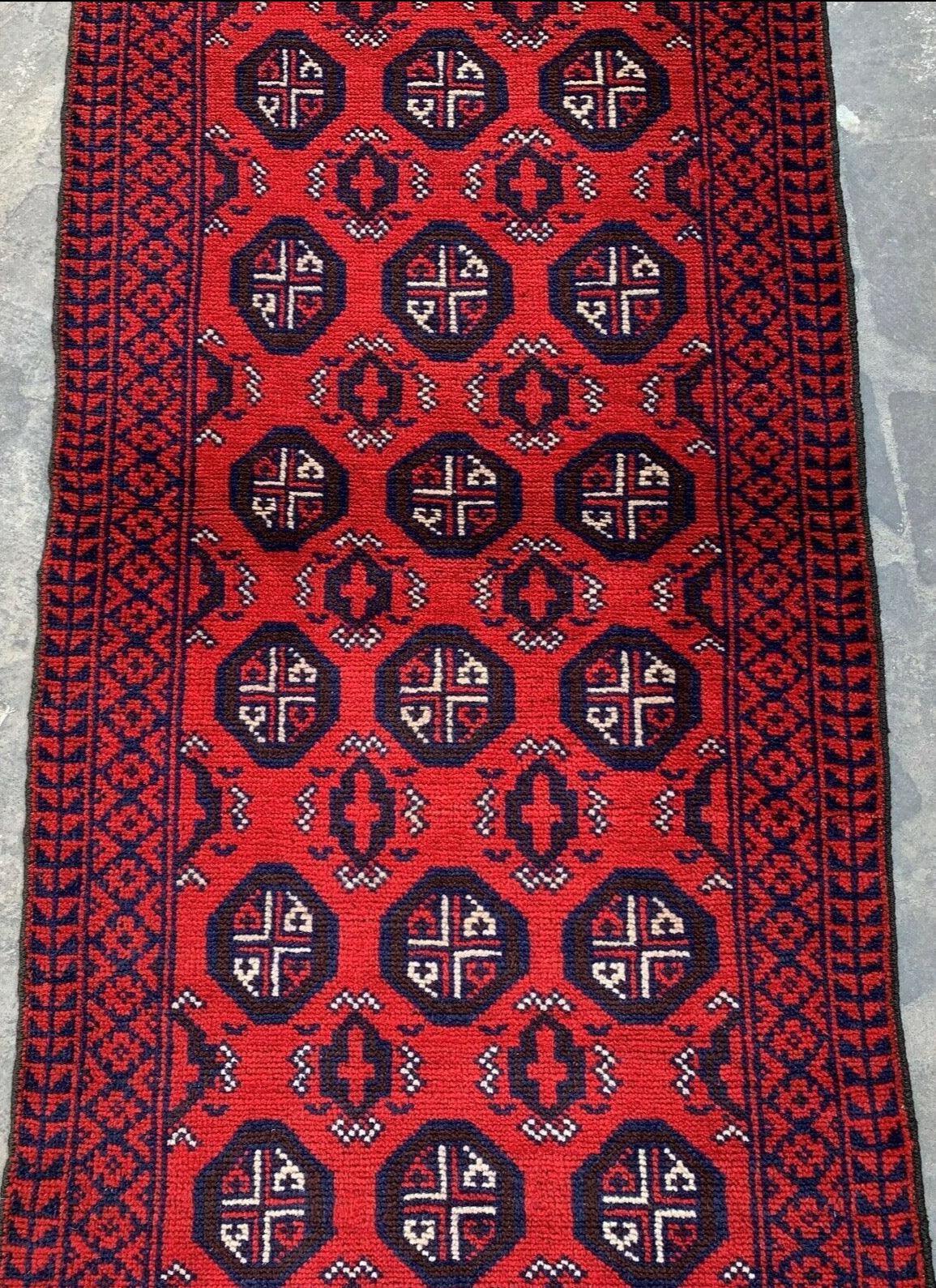 Hand knotted Afghani wool Baluch runner rug in red with intricate black geometric medallions and a border featuring repeating angular patterns, displayed on stone flooring.