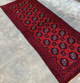 Hand-knotted Afghani Baluch runner rug in red with dark geometric medallions and intricate navy and black patterned borders, laid on a gray stone floor.