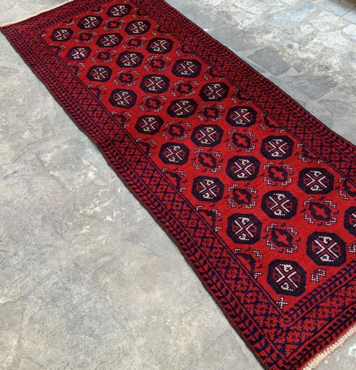 Hand-knotted Afghani Baluch runner rug in red with dark geometric medallions and intricate navy and black patterned borders, laid on a gray stone floor.