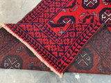 Close-up of a hand-knotted Afghani wool Baluch runner rug with red and dark blue geometric patterns and a fringed edge, partially folded to show the back.