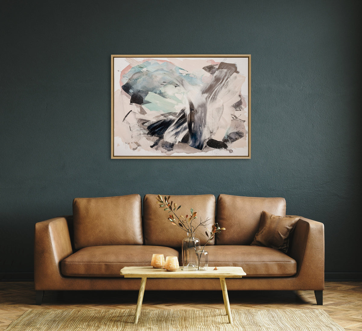 Abstract painting in soft blues, grays, blacks, and pinks framed in light wood, hanging above a tan leather sofa against a dark teal wall.