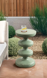 Sage green, glossy metal side table with three stacked rounded tiers supporting a flat circular top holding a glass of water with lemon slice in an outdoor garden setting.