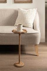 Small wooden drink table with scalloped edge and slender pedestal base holding a white ceramic mug, positioned in front of a beige upholstered sofa with a patterned pillow