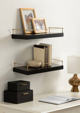 Two black floating shelves with thin gold metal railings mounted on a white wall, holding framed photos, books, and a small bowl, above a white desk with a gold lamp and black boxes.