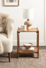 Compact wooden side table with a rounded top and lower shelf, holding a light wood vase lamp and white mug, placed beside a cream textured armchair with a fur throw.