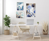Three framed abstract canvases with blue, beige, and black leaf-like patterns arranged on a white wall above a minimalist white desk and beige chair.