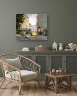 Painting of a yellow taxi on a city street with muted buildings and figures, displayed above a gray-green cabinet in a neutral-toned room with rattan chair and wood floor.