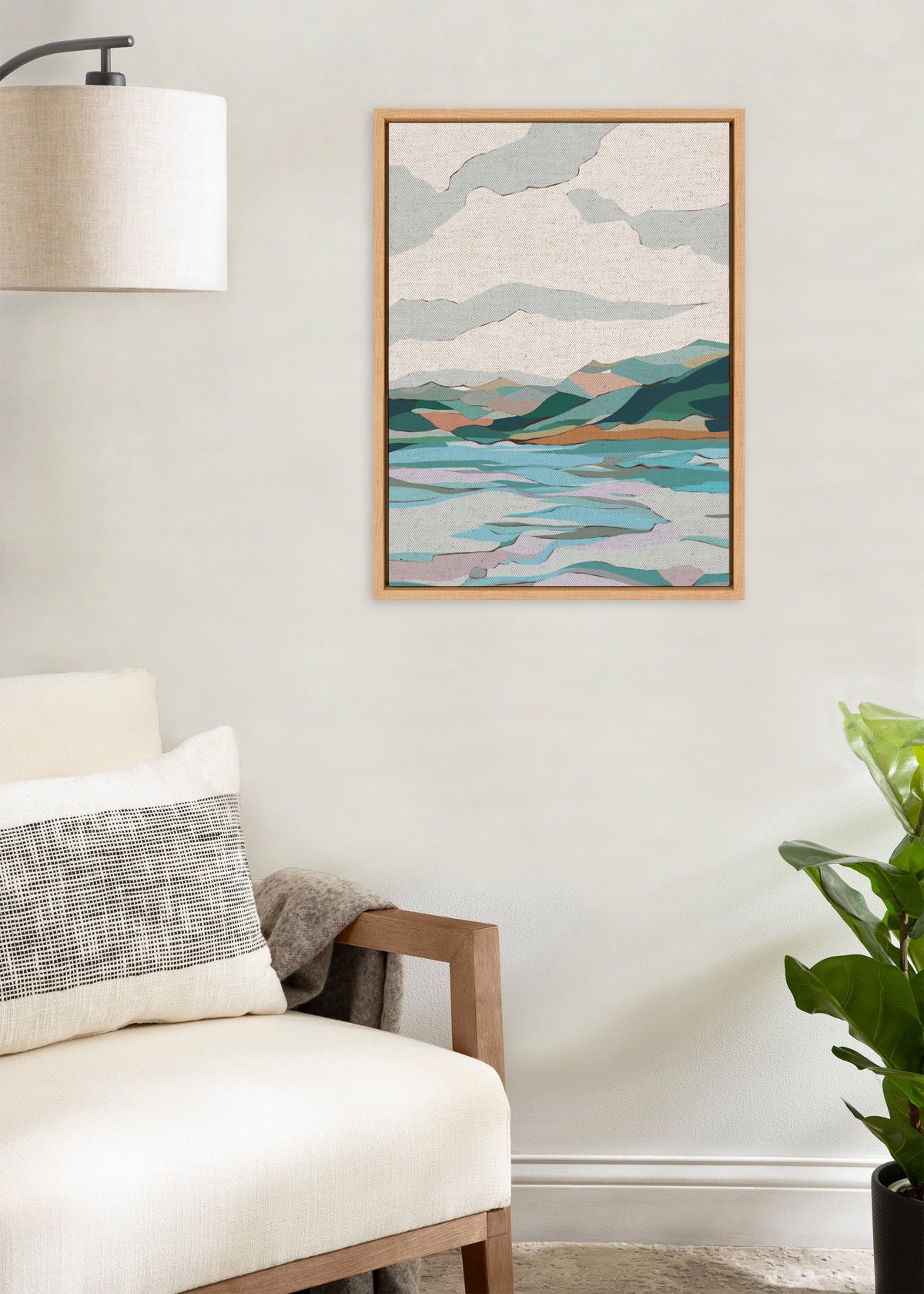 Framed canvas of stylized lake and mountain landscape with layered blue and green shapes under a cloudy beige sky, hanging on a light wall above a white chair.