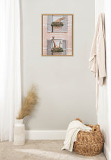 Framed canvas with two windows painted in muted pink and blue, each featuring giraffes peeking from flower-filled balconies, hung on a light gray wall above woven basket and vase.