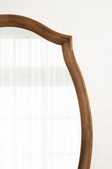 Close-up of a wooden scalloped wall mirror frame with a smooth warm brown finish against a white wall and sheer curtains reflected.