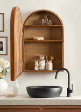 Wall-mounted wooden arch-shaped vanity cabinet with open door revealing two shelves holding glass jars and toiletries above a matte black round vessel sink and black faucet on a light countertop.