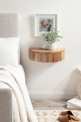 Round floating wood side table with natural wood grain mounted on white wall, holding a small white textured pot with green leafy plant and a pair of glasses nearby.
