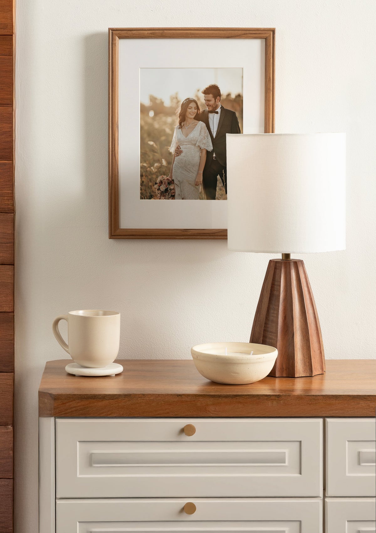 Wooden table topped with a geometric wooden table lamp with a white drum shade, a beige ceramic bowl, and a cream mug on a coaster, under a framed photo.