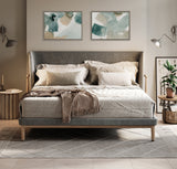 Gray upholstered bed with wooden legs and neutral bedding, flanked by two wall-mounted lamps and side tables with decor, beneath two abstract green and beige artworks.