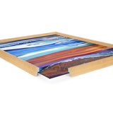 Framed art print featuring horizontal stripes in various shades of blue and reddish-brown, displayed with a light wood frame and a corner section removed revealing the print edge.
