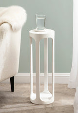 Tall, slender white side table with open, rounded rectangular cutouts serves as a pedestal for a glass of water, set beside a cream textured armchair.
