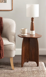 Small wooden side table with rounded triangular top and curved legs, holding a white mug and a white lamp with a wooden base, next to a beige upholstered armchair.