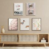 Five framed art prints arranged in two rows over a woven panel wooden sideboard, featuring a running dog with colorful circles, a giraffe peeking from window boxes, pink Paris streets, dogs in striped shirts, and soft pink flowers