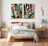 Modern bedroom with wooden bed and white bedding featuring two framed abstract leaf-patterned canvases in teal, orange, black, and pink hues on a white wall.