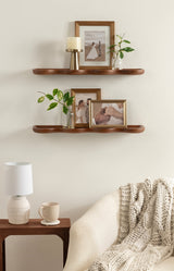 Two smooth, warm brown wooden shelves mounted on a light beige wall holding framed photos, a glass vase with green branches, and a small brass candle holder with a white candle.