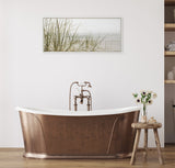 Copper freestanding bathtub with antique-style faucet, positioned in front of a white wall with a framed calming beach grass canvas and a rustic wooden stool holding flowers.