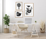 Minimalist workspace with a light wood trestle desk, beige chair, and three black and white linocut art prints featuring stylized birds and a tree in wooden frames.