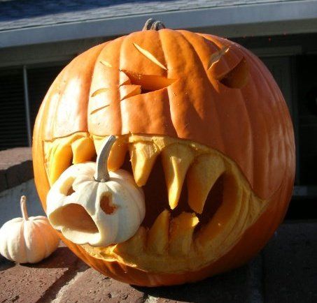 Orange pumpkin carved with angry eyes and jagged teeth biting down on a smaller white pumpkin held in its mouth, displayed outdoors on a brick surface.