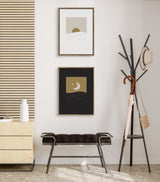 Two framed minimalist canvas art pieces on a wall, one with a gold sun and light gray background, the other with a gold crescent moon on black, above a dark bench and beside a black coat rack.