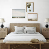 Minimalist bedroom with light wood bed and matching nightstands, white linens, black lampshades, and three framed nature-themed artworks on the white wall.