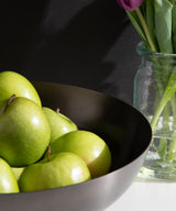 Dark ribbed metal bowl filled with green apples, positioned next to a glass vase holding pink tulips with green leaves on a white surface.