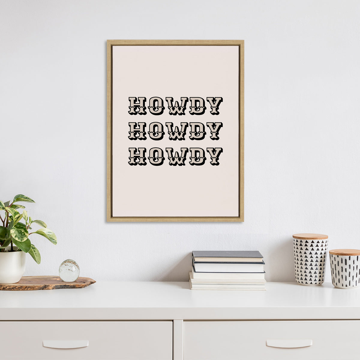 Wall art with the word 'HOWDY' repeated three times in bold western-style black lettering on a light pink background, framed in simple natural wood, hanging above a white desk with books, plants, and containers.