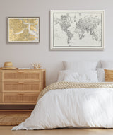 Bedroom with white bedding and beige knitted throw, light wood dresser with hat and coral decor, framed black-and-white world map and yellow-toned vintage map on the light beige wall.