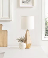 Geometric wooden table lamp base with a white cylindrical shade, placed beside a small round white vase with dried flowers on a white surface near a window.