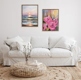 Two framed paintings above a white sofa: left shows a sunset over ocean waves with pink and blue hues, right displays pink flowers with green leaves and blurred background.