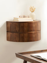 Curved floating wooden side table with smooth grain, holding stacked books, glasses, and a textured vase with dried wheat, against a soft cream wall.
